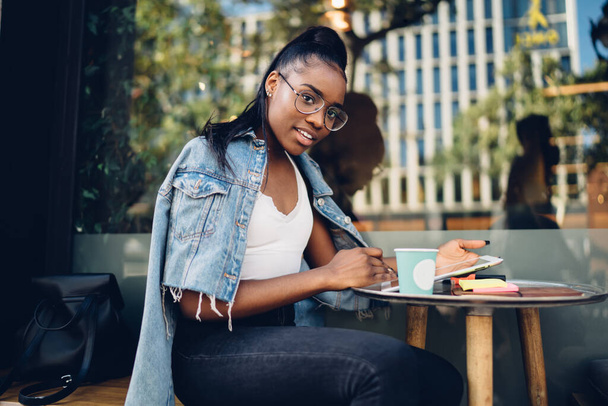 Side view of smiling African American stylish lady in glasses sitting at table by glass wall with cup of coffee and surfing tablet while looking at camera - Фото, зображення