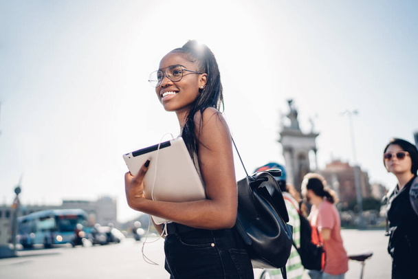 From below side view of positive young black woman in back lit holding tablet and earphones standing on roadside of downtown - Fotografie, Obrázek