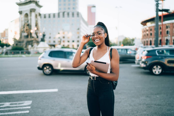 Happy young African American woman in trendy wear and eyeglasses standing on road with tablet and smiling at camera while enjoying summer sunny day - Fotografie, Obrázek