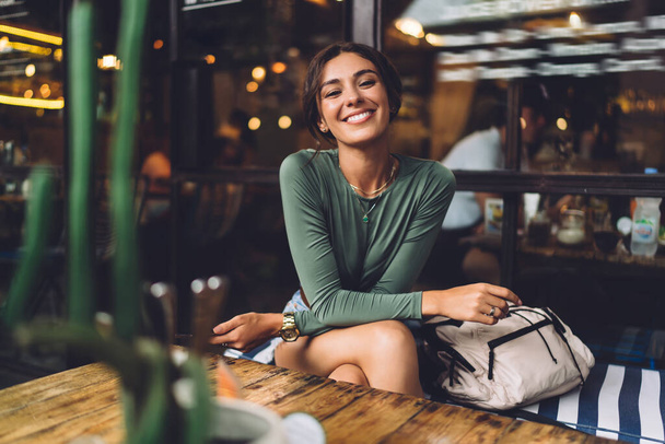 Wesoła kobieta w ubraniu casual siedzi przy drewnianym stole i uśmiecha się patrząc w kamerę i trzymając smartfona w ręku - Zdjęcie, obraz