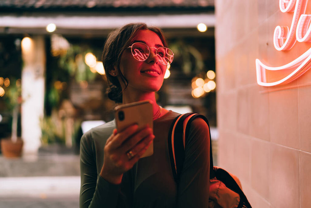 Modern content female in casual outfit and eyeglasses with backpack smiling and looking away while browsing smartphone on street near neon sign - Φωτογραφία, εικόνα