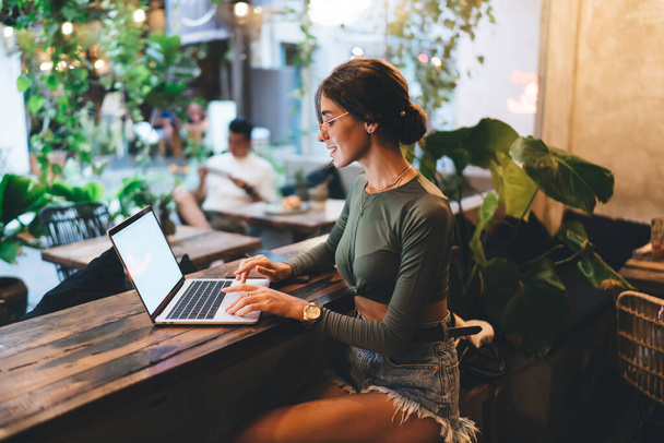 Υψηλής γωνίας πλαϊνή όψη θετικής γυναίκας ελεύθερου επαγγελματία σε casual wear και γυαλιά που κάθεται στο τραπέζι με laptop και χαμογελώντας - Φωτογραφία, εικόνα