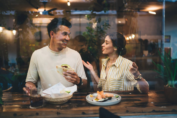 Diverse young couple in love in casual outfit having pleasant conversation and enjoying meal together while sitting at bar counter in cafe behind glass window - Фото, зображення
