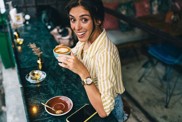 From above smiling woman in trendy clothes drinking cup of hot coffee while relaxing at table with decor item outside cozy restaurant - Fotografie, Obrázek