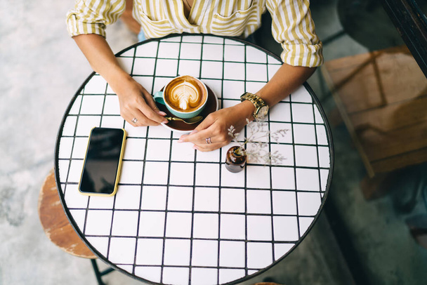 From above of anonymous female holding cup of fresh cappuccino with froth while resting at round table with smartphone - 写真・画像