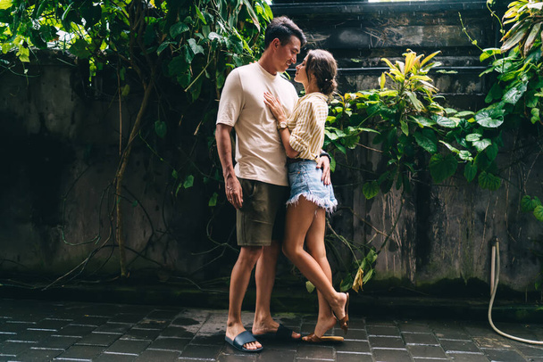 Full body of couple in love standing on pavement near shabby wall decorated with tropical foliage and looking at each other - Fotó, kép