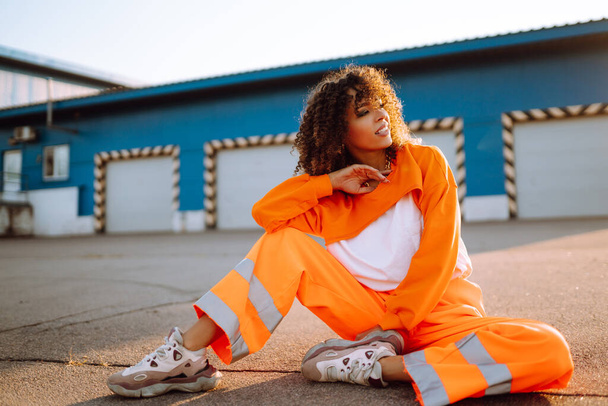 Fashion sporty woman in an orange suit posing in the street at sunset. Stylish woman with curly hair. Lifestyle, sport,  youth concept. - Foto, imagen