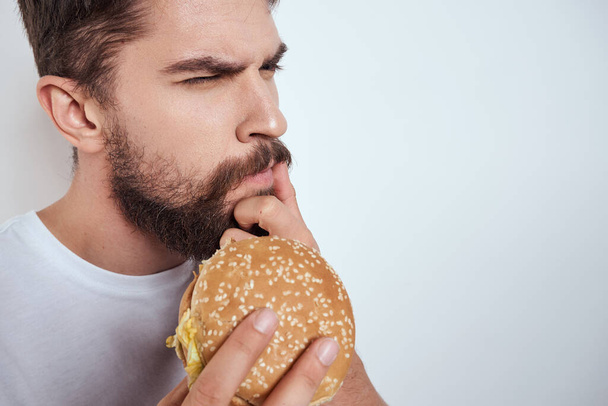 Ένας άνθρωπος τρώει ένα χάμπουργκερ σε ένα ελαφρύ φόντο σε ένα λευκό T-shirt crop view close-up πείνα fast food - Φωτογραφία, εικόνα