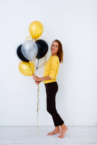 Full-length portrait of a young woman in a yellow sweater holding a bunch of balloons in gold, white and silver against a white wall. - 写真・画像