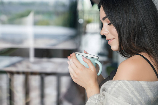 Uśmiechnięta brunetka w domu pokryta kratą pijąca poranną kawę przy oknie. - Zdjęcie, obraz