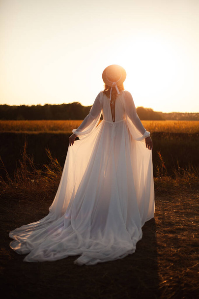 Happy woman standing with her back on sunset in nature . Woman in a long white dress with a hat on her head - Zdjęcie, obraz