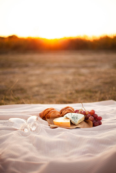 Romanttinen piknik kahdelle auringonlaskun aikaan. Peitossa on kaksi juustonpalaa, viinirypäleitä, kaksi croissantia ja kaksi tyhjää lasia. Lähikuva valokuva - Valokuva, kuva