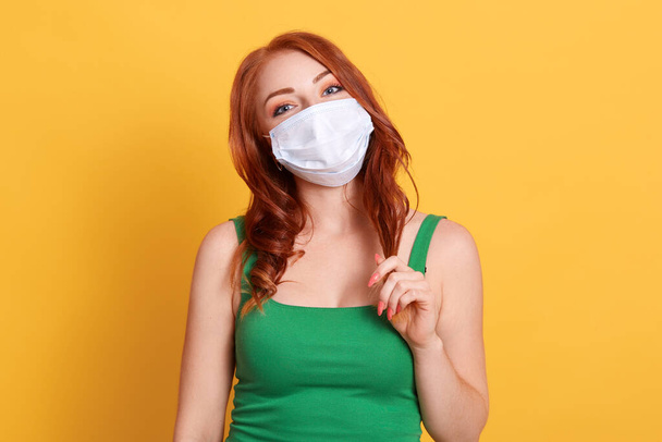 Close up shot of young woman with wavy hair, wears green t shit and medical disposable mask, cares about her health, protects in dangerous situation, standing against yellow wall. - Foto, imagen
