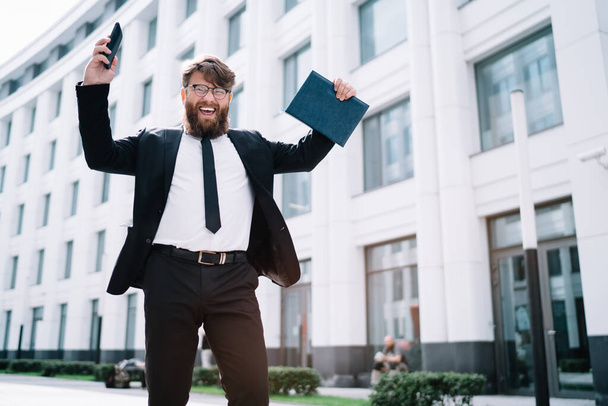 Low angle of cheerful bearded businessman in formal wear and eyeglasses standing near modern building and celebrating success with smartphone and notebook in hands - 写真・画像