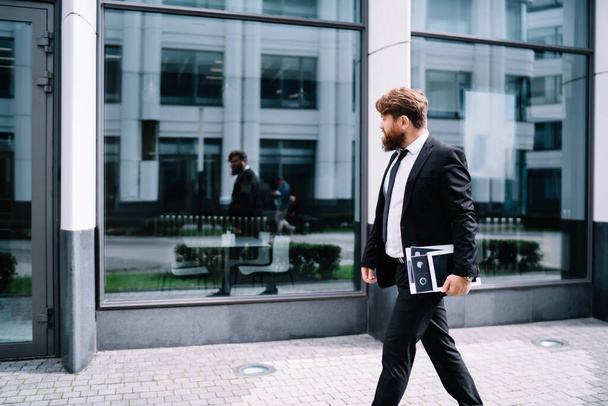 Side view of serious bearded businessman in formal clothes and tie walking near modern building and looking away while holding documents - Fotografie, Obrázek