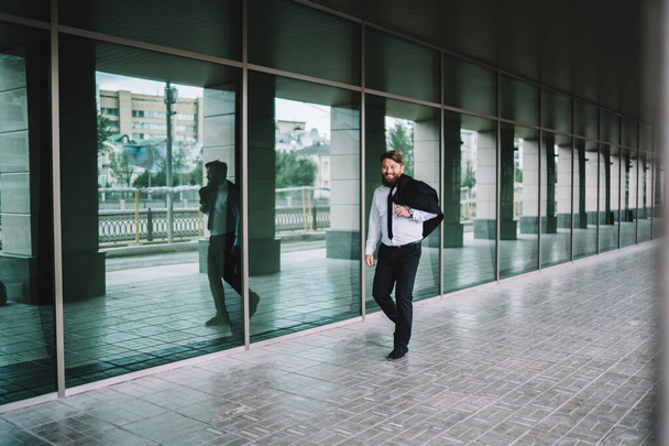 Full body of positive entrepreneur with bushy beard in trendy formal wear walking along modern glass building and laughing while looking at camera - 写真・画像