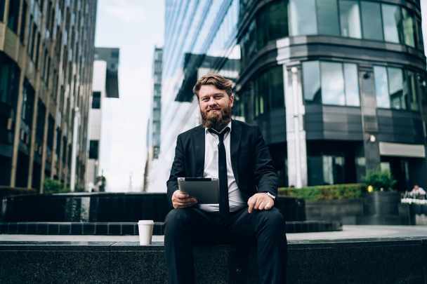 Low angle of elegant bearded male manager in formal clothes and glasses sitting on stone border on street and looking at camera while working on tablet - Photo, Image