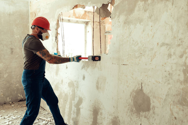 Elinde çekiçle inşaatçı beton duvarı yıkar inşaatçı koruyucu bir kostüm ve kask giyer. - Fotoğraf, Görsel