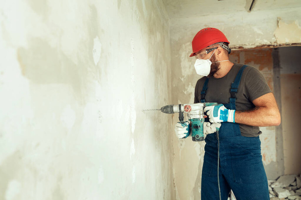 строитель с перфоратором сверлит отверстия в бетонной стене строитель одет в защитный костюм и шлем - Фото, изображение