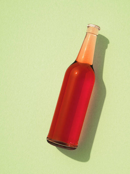 One transparent glass bottle with metal cap laying on light green surface. Beer bottle without label, red liquid inside. Retro drink bottle concept. - Fotoğraf, Görsel