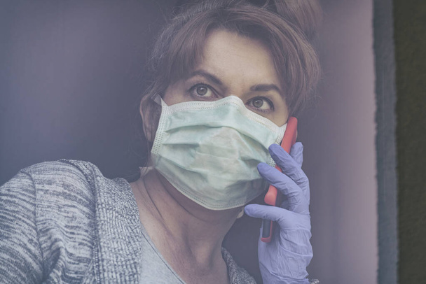 Nainen, jolla on suojaava kasvonaamio ja joka katsoo ulos kotinsa ikkunasta koronaviruksen covid-19-epidemian vuoksi. Kotikaranteeni ja eristäytyminen - Valokuva, kuva