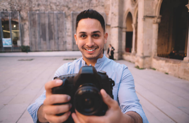 Happy ethnic tourist in casual shirt and professional photo camera standing on street of historic town and smiling while looking at camera - Zdjęcie, obraz