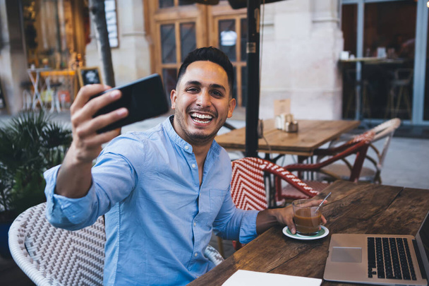 Young positive ethnic man in stylish light blue shirt talking via video link while having break and drinking tea in street cafe and looking at camera with toothy smile - Фото, изображение