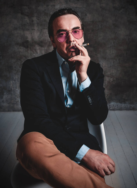 Brutal man with slicked back hair in official suit and smoked blue glasses sits with his legs crossed smoking cigarette - Foto, afbeelding