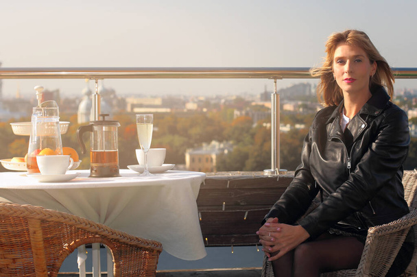una hermosa mujer rusa de mediana edad se sienta en una mesa en un restaurante en la azotea con un panorama de la ciudad de otoño a la luz de la brillante puesta de sol - Foto, imagen