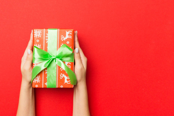 Vrouwelijke handen met gestreepte geschenkdoos met gekleurd lint op rode achtergrond. Kerstconcept of andere vakantie handgemaakte geschenkdoos, concept bovenaanzicht met kopieerruimte. - Foto, afbeelding