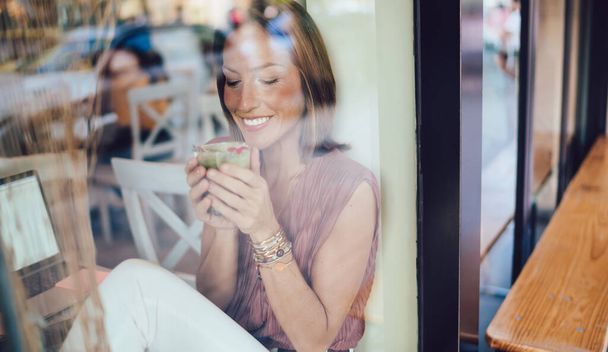 Door een glas vrolijke vrouw in stijlvolle outfit glimlachend en genietend van een glas frisse cocktail terwijl ze met laptop in café zit - Foto, afbeelding