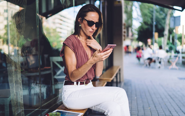 Serious middle aged woman in trendy wear and sunglasses looking pensively on mobile phone and touching chin while sitting on street and surfing internet - Photo, Image
