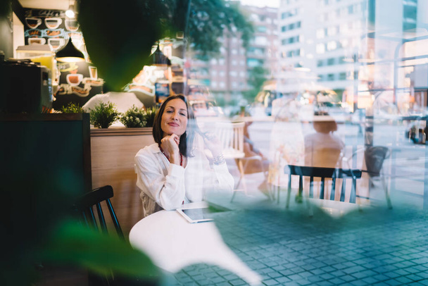 Cheerful mature woman in trendy clothes sitting at table with tablet and touching hair while resting through window in modern cafe - Foto, Imagem