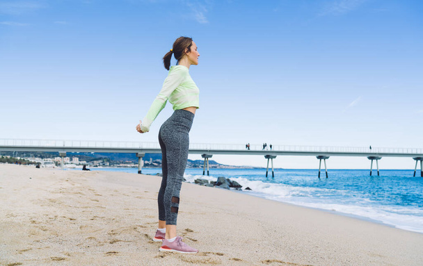Strong female trainer in tracksuit for morning jogging stretching body muscles during warm up exercising at coastline,flexible fit girl enjoying outdoors pilates during leisure time at serene seashore - Foto, Imagem
