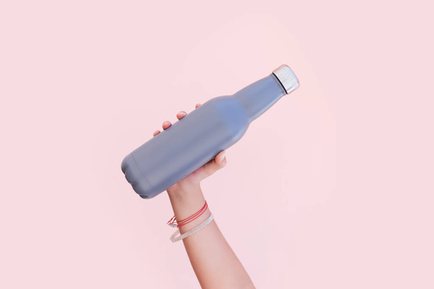 Közelkép a női kéz kezében egy öko-újrahasznosítható acél rozsdamentes termo vizes palack a háttérben pasztell rózsaszín színű. - Fotó, kép