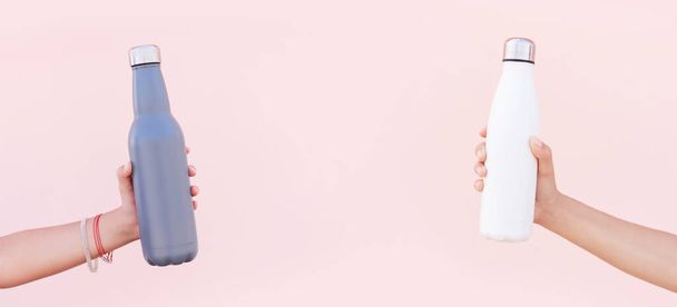 Крупный план женщин руки держа eco многоразовой стали из нержавеющей термоводы бутылки белого и синего цветов, на фоне пастельно-розового цвета. - Фото, изображение