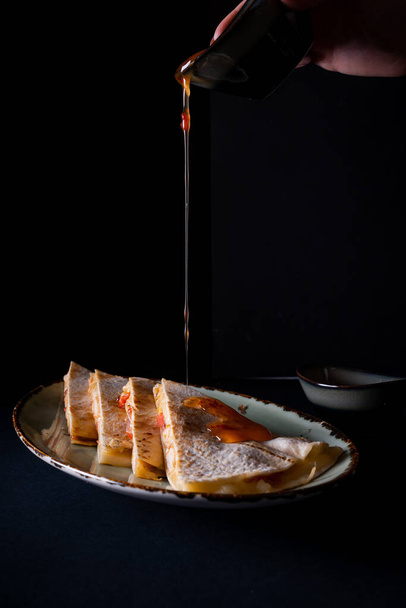 A vertical shot of female hand pouring a sauce into appetizing chicken quesadilla on black background - Foto, afbeelding