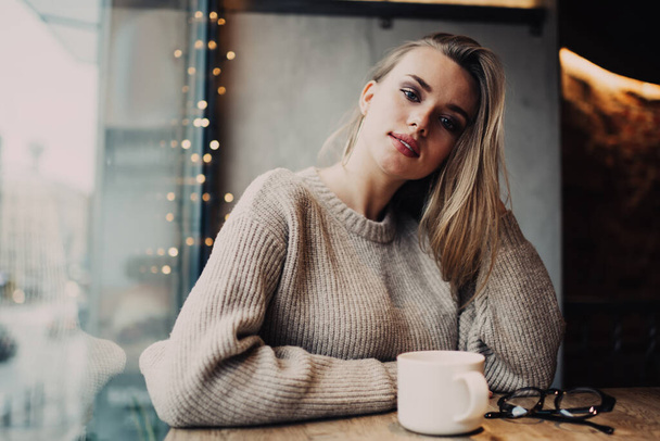 Retrato de atractiva cliente femenina disfrutando de tiempo de fin de semana para visitar la cafetería con cappuccino aroma para el calentamiento, hermosa chica hipster caucásica 20 años con taza de té mirando a la cámara - Foto, Imagen
