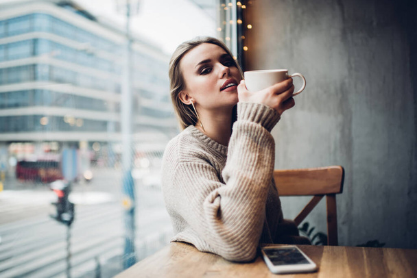 Attractive female customer with caffeine beverage in hand sitting near cafeteria window and looking at camera, charming hipster girl 20s holding hot tea for warming and posing in cafe interior - Foto, Imagem