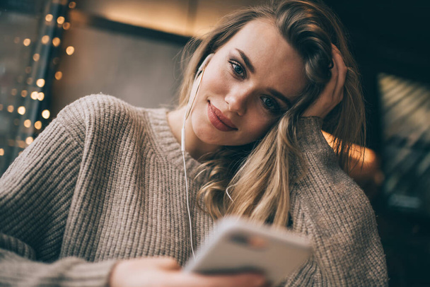 Low angle of young cheerful female with long blond hair in knitted sweater with light smile looking at camera while listening to music - Zdjęcie, obraz
