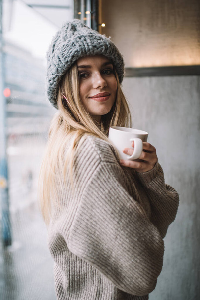 Half length portrait of trendy dressed customer posing near cafeteria window enjoying coffee time, carefree female in knitted pullover and winter hat holding aroma caffeine beverage for warming - Foto, Imagem