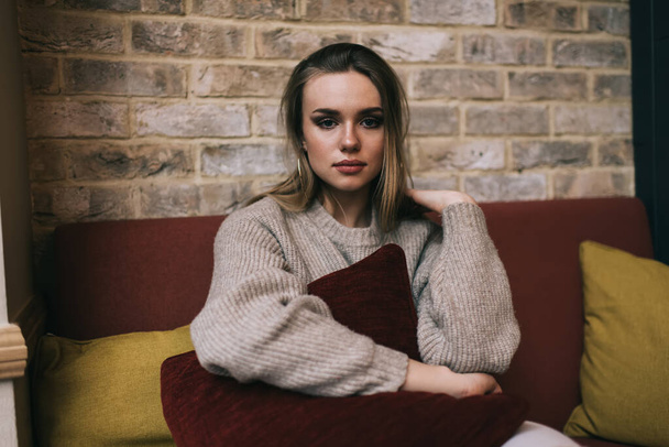 Portrait of Caucasian hipster girl with pillow in hands resting at comfortable sofa, female student dressed in knitted pullover looking at camera during weekend recreation in loft cafeteria interior - Fotografie, Obrázek