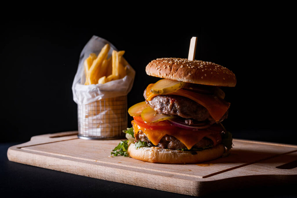 Un primer plano de una hamburguesa doble de ternera y papas fritas en un tablero de madera sobre un fondo negro - Foto, Imagen
