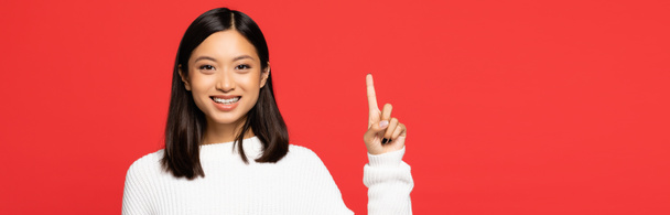 happy asian woman in sweater pointing with finger isolated on red, banner - Fotó, kép
