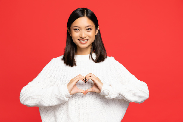 happy and young asian woman showing heart sign with hands isolated on red - 写真・画像
