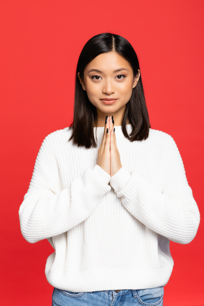 happy and young asian woman standing with praying hands isolated on red - Foto, immagini