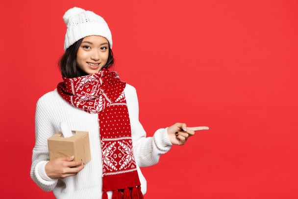счастливая и молодая азиатская женщина в шляпе и шарфе держа ткань коробки и указывая пальцем изолированы на красный  - Фото, изображение