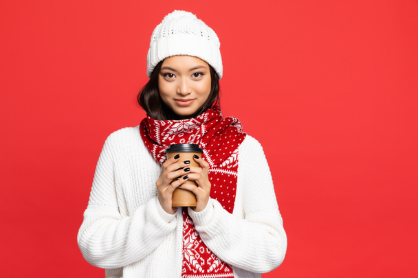 mujer asiática feliz en sombrero y bufanda sosteniendo taza desechable con café para ir aislado en rojo - Foto, Imagen