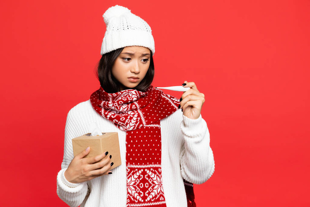 帽子とスカーフの病気のアジア人女性は赤で隔離された電子温度計とティッシュボックスを持っています  - 写真・画像