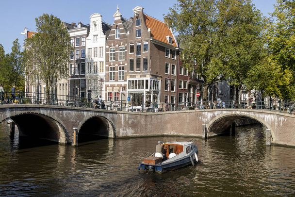 AMSTERDAM, PAESI BASSI - 22 / 09 / 2020: Barca con bandiera locale di Amsterdam e persone provenienti da sotto un ponte che passano davanti alle facciate iconiche ed esterne delle dimore olandesi sul canale - Foto, immagini